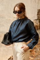 A woman stands in front of a textured wall, wearing a dark blue Gathered Collar Metallic Top by Maison Lilli and light beige high-waisted trousers. She holds a black clutch and wears dark sunglasses, her hair tied back. The subtle sheen of the blouse's metallic fabric adds an extra touch of glamour to her ensemble.