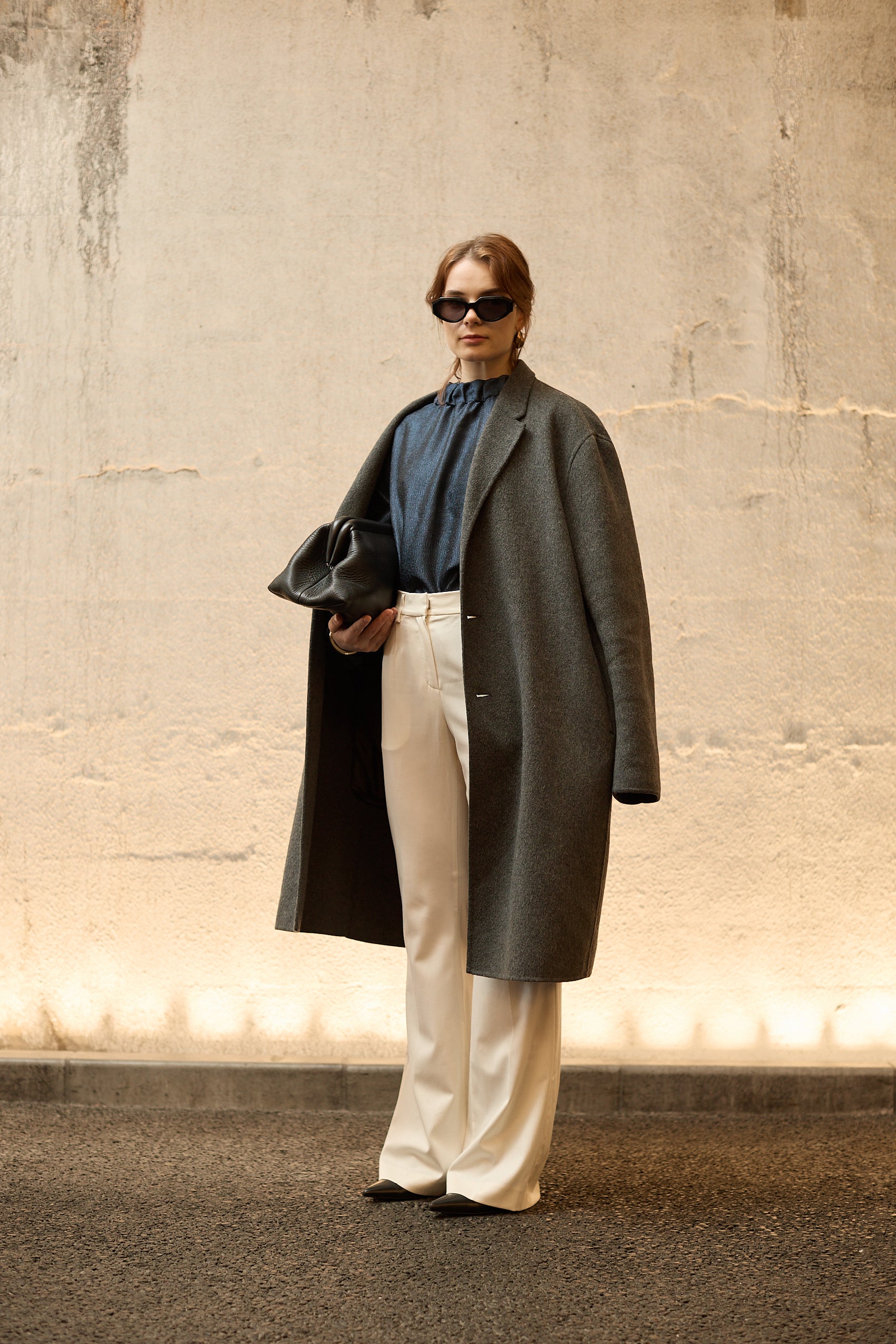 A woman stands in front of a textured wall, wearing a dark blue Gathered Collar Metallic Top by Maison Lilli and light beige high-waisted trousers. She holds a black clutch and wears dark sunglasses, her hair tied back. The subtle sheen of the blouse's metallic fabric adds an extra touch of glamour to her ensemble.