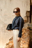 A woman stands in front of a textured wall, wearing a dark blue Gathered Collar Metallic Top by Maison Lilli and light beige high-waisted trousers. She holds a black clutch and wears dark sunglasses, her hair tied back. The subtle sheen of the blouse's metallic fabric adds an extra touch of glamour to her ensemble.