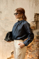 A woman stands in front of a textured wall, wearing a dark blue Gathered Collar Metallic Top by Maison Lilli and light beige high-waisted trousers. She holds a black clutch and wears dark sunglasses, her hair tied back. The subtle sheen of the blouse's metallic fabric adds an extra touch of glamour to her ensemble.