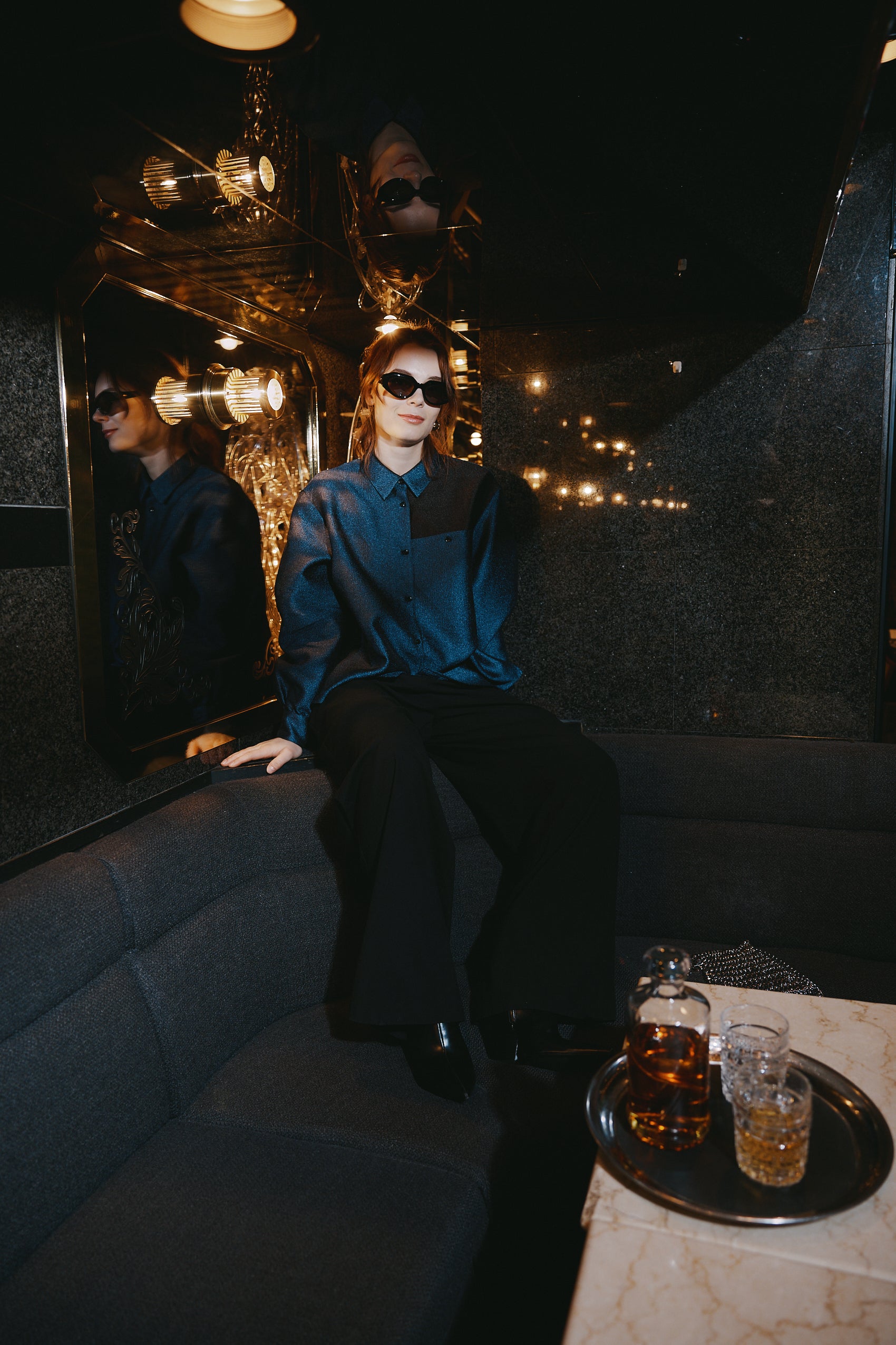 A woman stands in front of a textured wall, wearing a dark blue Gathered Collar Metallic Top by Maison Lilli and light beige high-waisted trousers. She holds a black clutch and wears dark sunglasses, her hair tied back. The subtle sheen of the blouse's metallic fabric adds an extra touch of glamour to her ensemble.