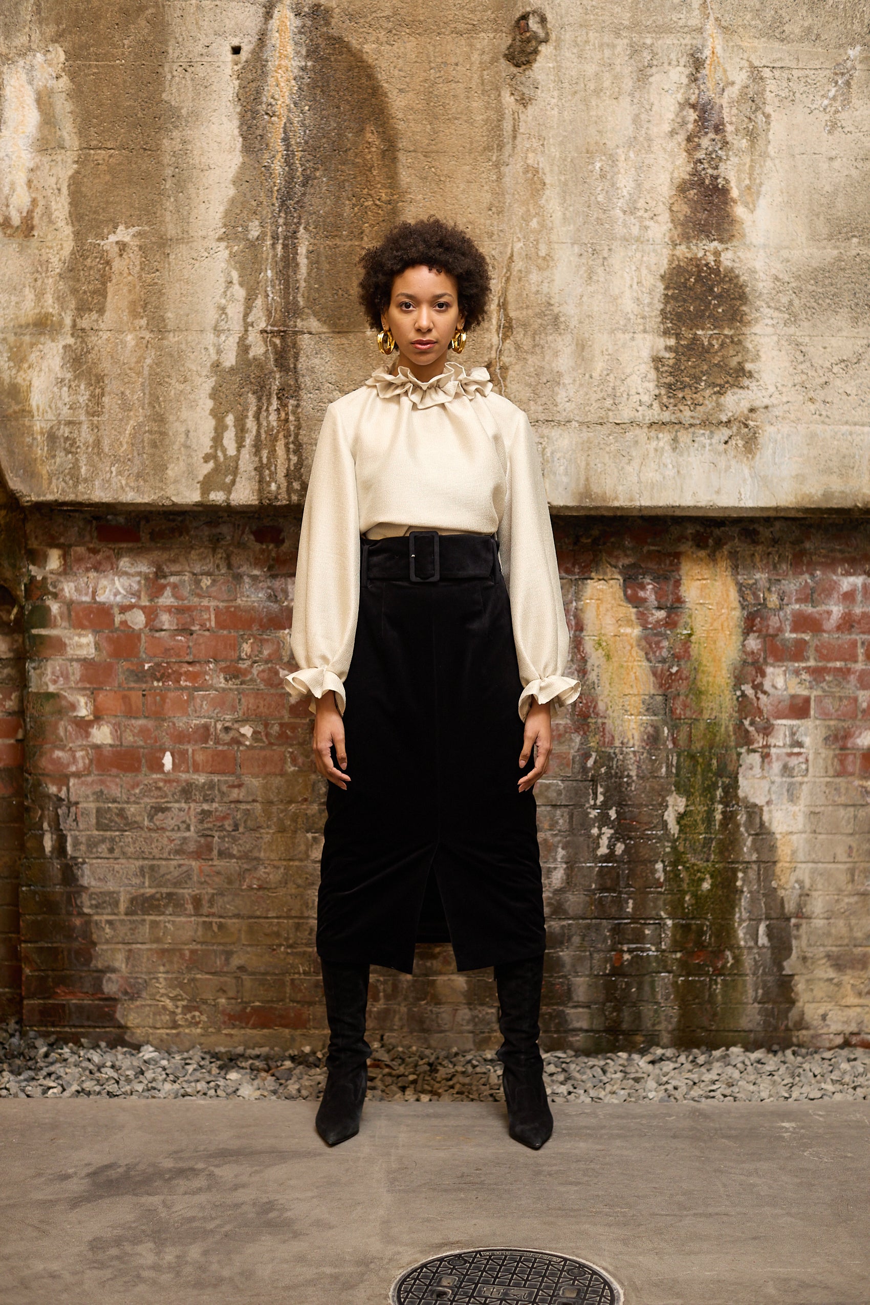 A woman stands in front of a textured wall, wearing a dark blue Gathered Collar Metallic Top by Maison Lilli and light beige high-waisted trousers. She holds a black clutch and wears dark sunglasses, her hair tied back. The subtle sheen of the blouse's metallic fabric adds an extra touch of glamour to her ensemble.