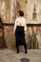 A woman stands in front of a textured wall, wearing a dark blue Gathered Collar Metallic Top by Maison Lilli and light beige high-waisted trousers. She holds a black clutch and wears dark sunglasses, her hair tied back. The subtle sheen of the blouse's metallic fabric adds an extra touch of glamour to her ensemble.