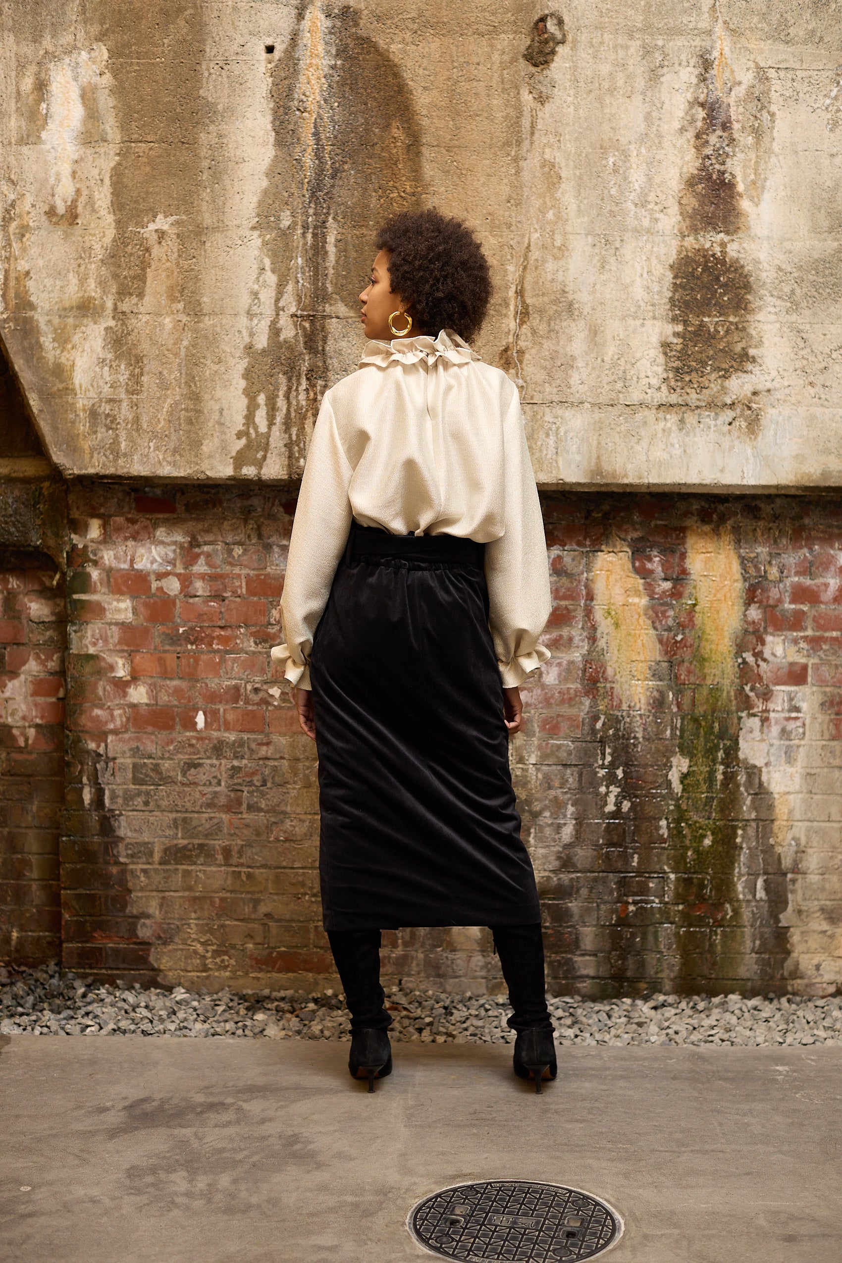 A woman stands in front of a textured wall, wearing a dark blue Gathered Collar Metallic Top by Maison Lilli and light beige high-waisted trousers. She holds a black clutch and wears dark sunglasses, her hair tied back. The subtle sheen of the blouse's metallic fabric adds an extra touch of glamour to her ensemble.