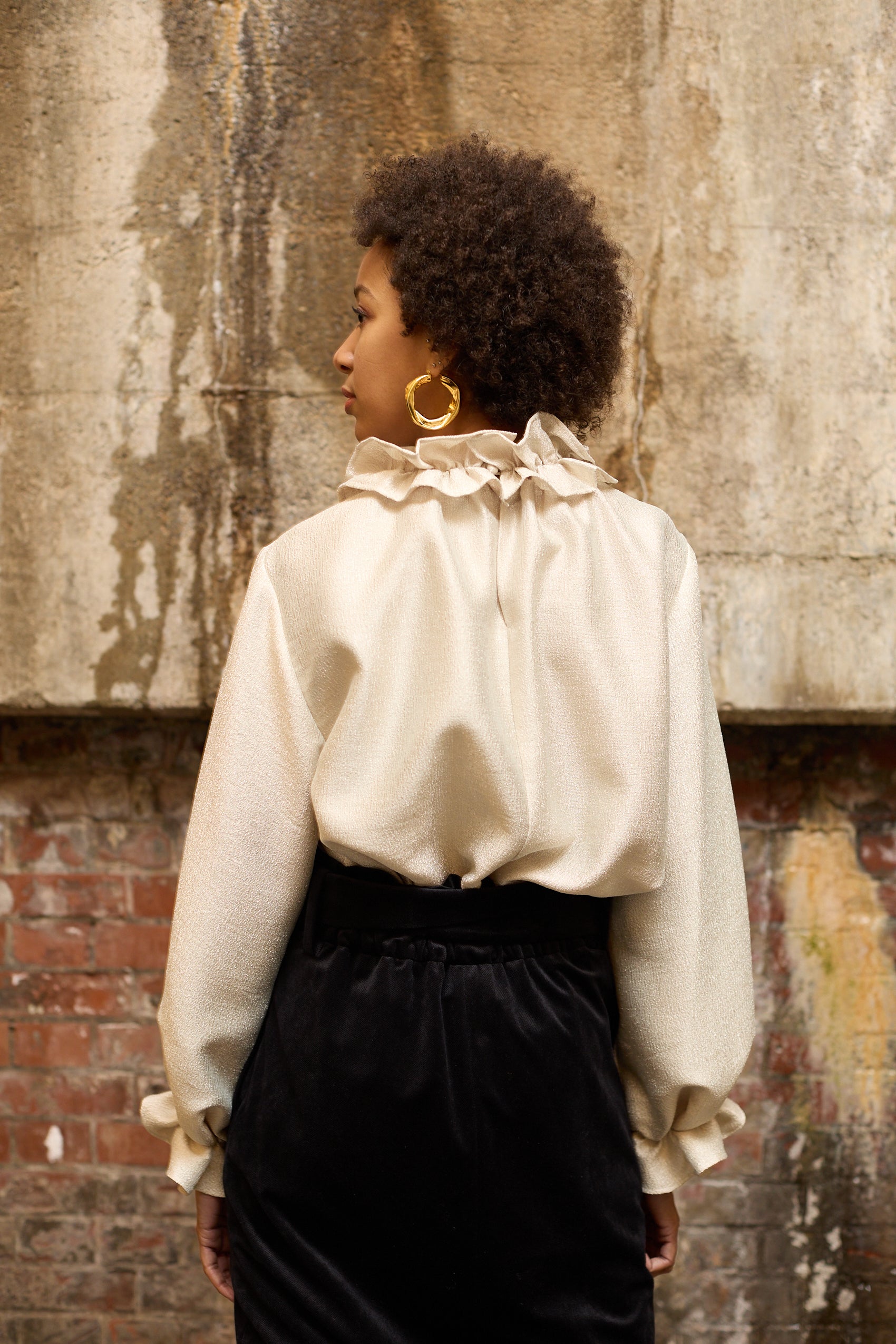 A woman stands in front of a textured wall, wearing a dark blue Gathered Collar Metallic Top by Maison Lilli and light beige high-waisted trousers. She holds a black clutch and wears dark sunglasses, her hair tied back. The subtle sheen of the blouse's metallic fabric adds an extra touch of glamour to her ensemble.