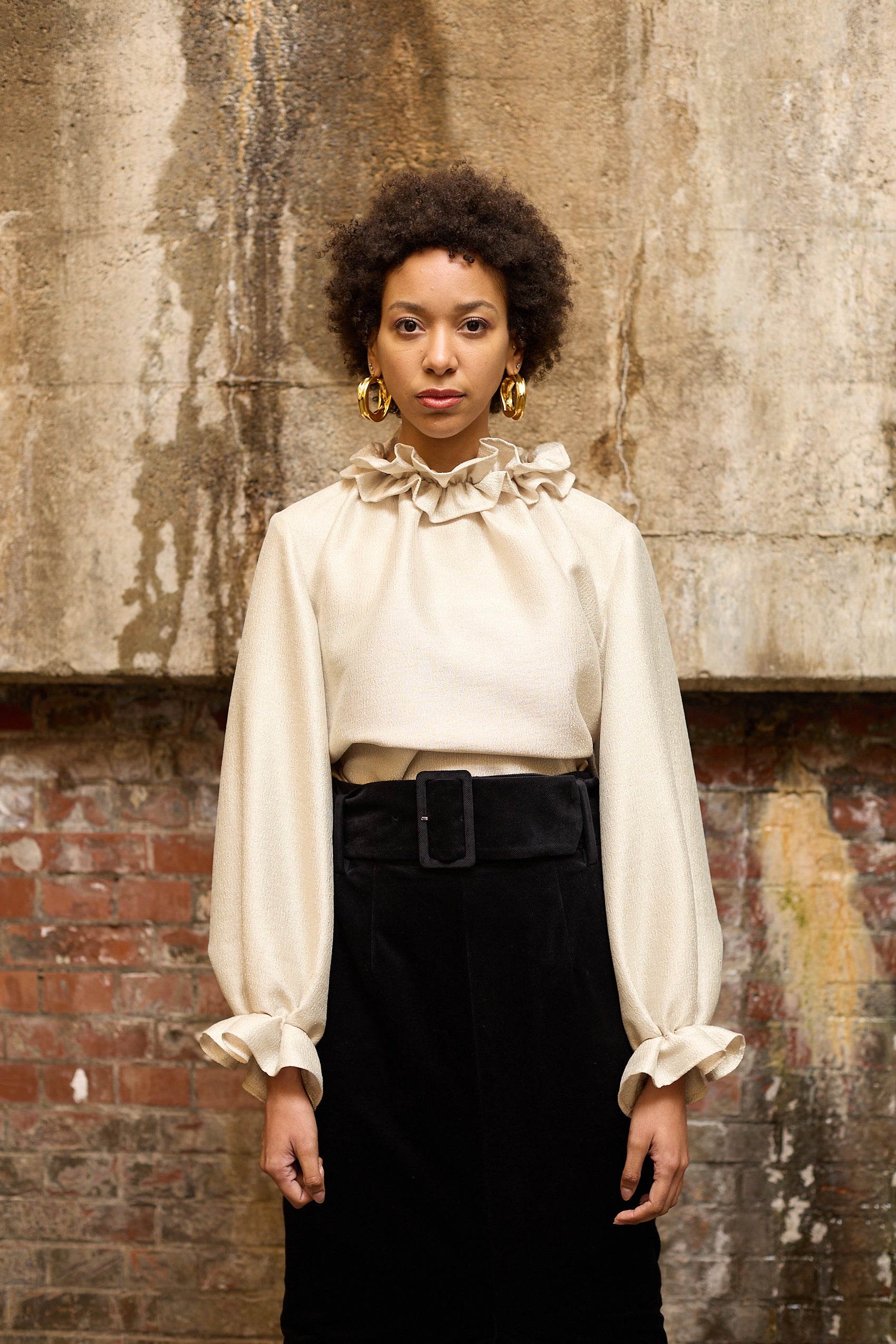 A woman stands in front of a textured wall, wearing a dark blue Gathered Collar Metallic Top by Maison Lilli and light beige high-waisted trousers. She holds a black clutch and wears dark sunglasses, her hair tied back. The subtle sheen of the blouse's metallic fabric adds an extra touch of glamour to her ensemble.