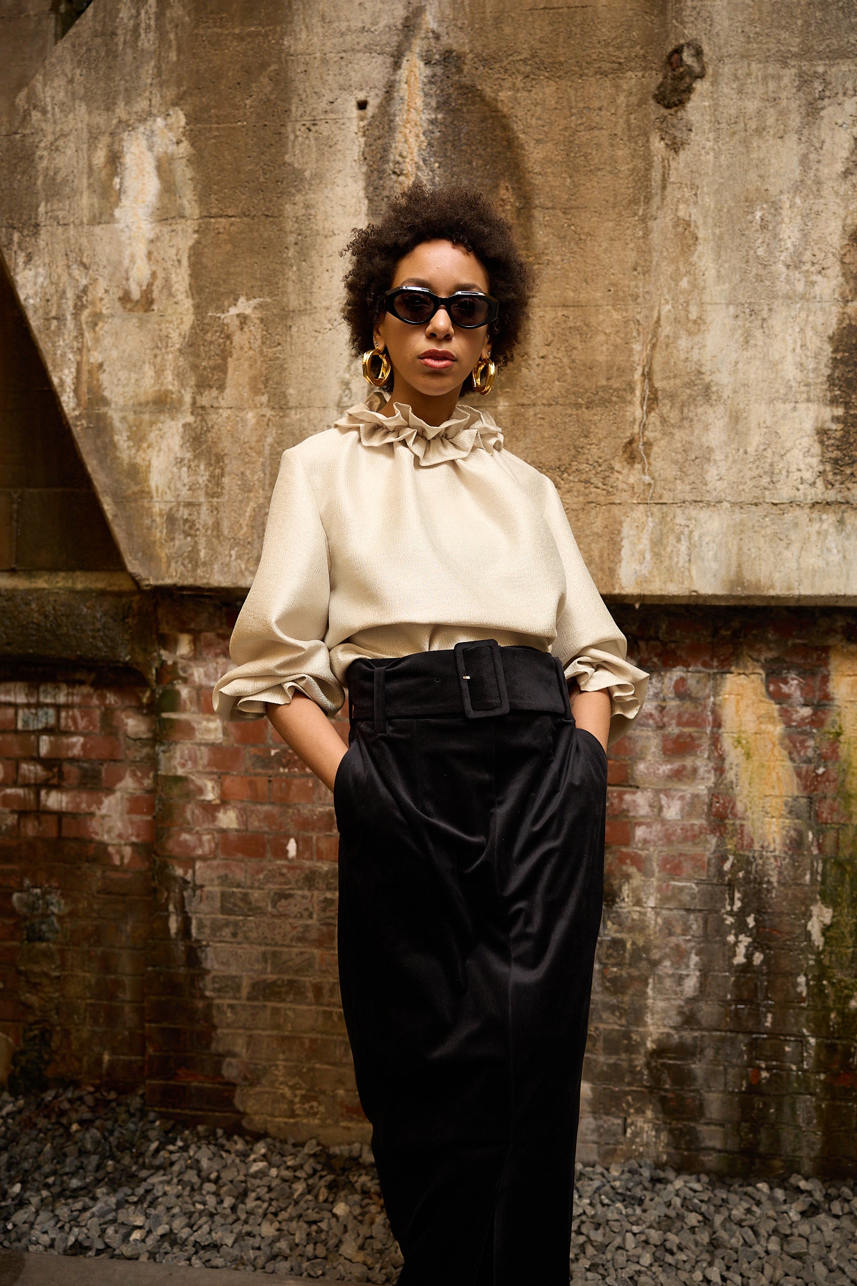 A woman stands in front of a textured wall, wearing a dark blue Gathered Collar Metallic Top by Maison Lilli and light beige high-waisted trousers. She holds a black clutch and wears dark sunglasses, her hair tied back. The subtle sheen of the blouse's metallic fabric adds an extra touch of glamour to her ensemble.