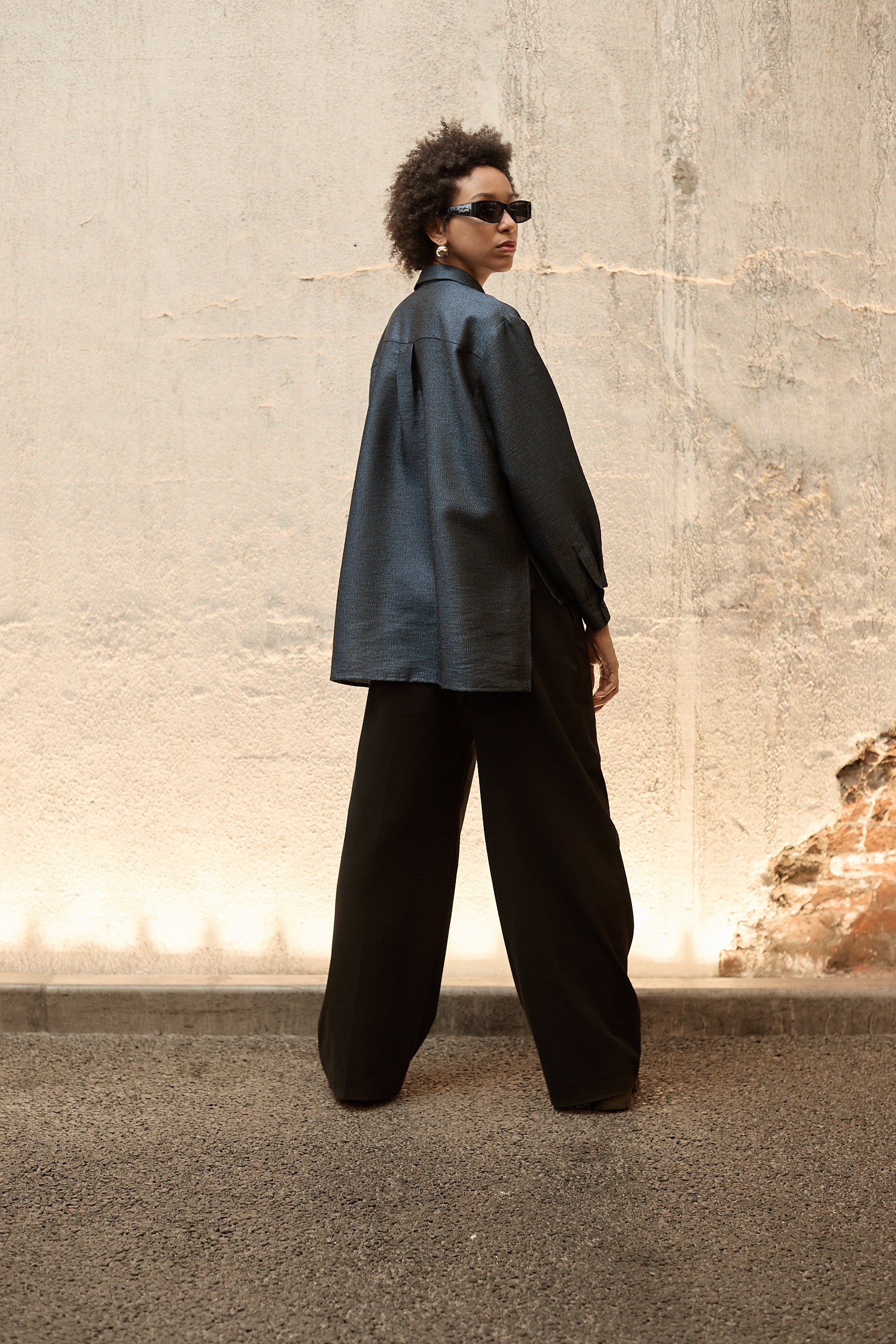 A woman stands in front of a textured wall, wearing a dark blue Gathered Collar Metallic Top by Maison Lilli and light beige high-waisted trousers. She holds a black clutch and wears dark sunglasses, her hair tied back. The subtle sheen of the blouse's metallic fabric adds an extra touch of glamour to her ensemble.