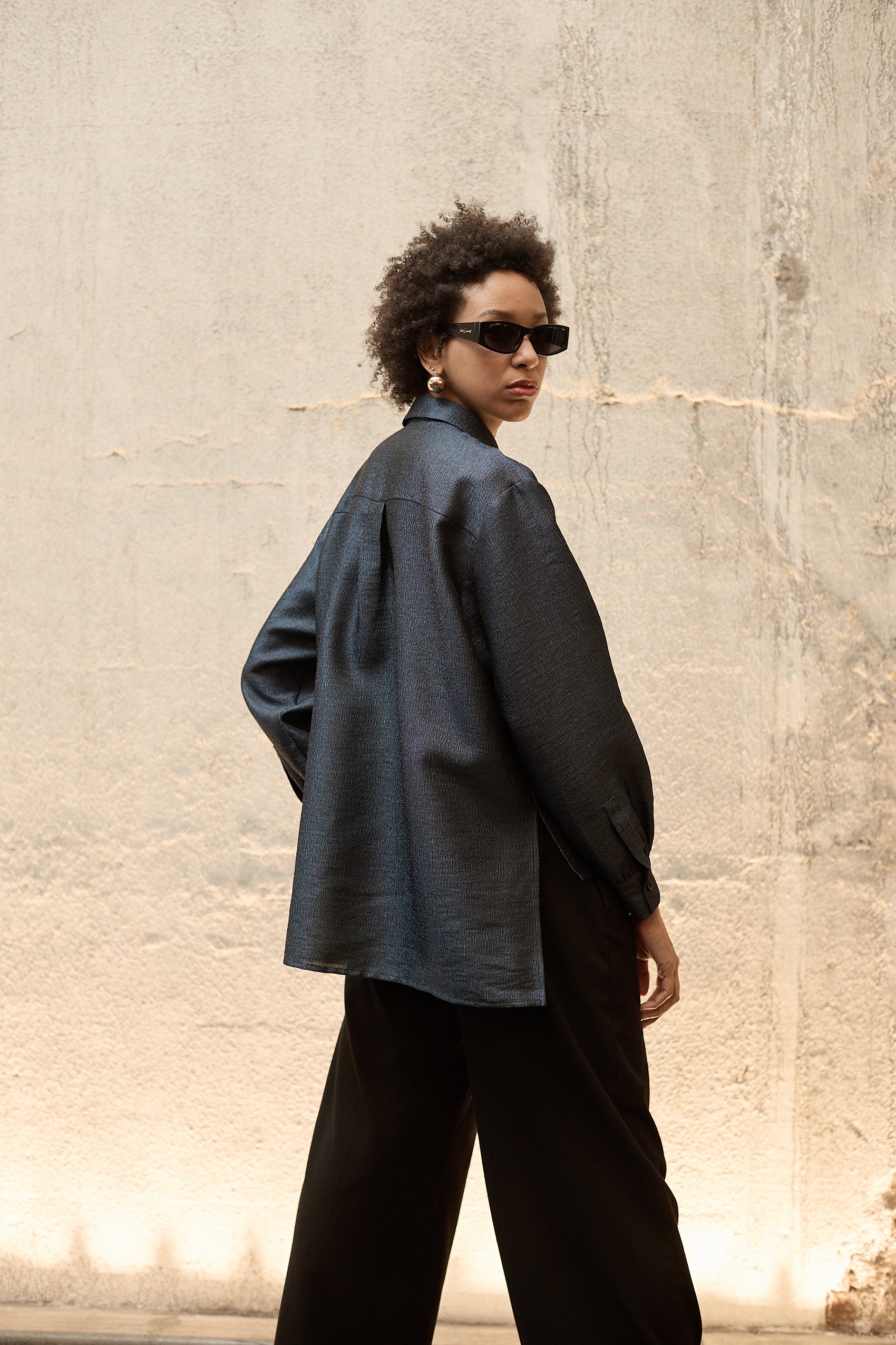 A woman stands in front of a textured wall, wearing a dark blue Gathered Collar Metallic Top by Maison Lilli and light beige high-waisted trousers. She holds a black clutch and wears dark sunglasses, her hair tied back. The subtle sheen of the blouse's metallic fabric adds an extra touch of glamour to her ensemble.