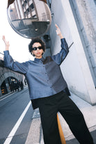 A woman stands in front of a textured wall, wearing a dark blue Gathered Collar Metallic Top by Maison Lilli and light beige high-waisted trousers. She holds a black clutch and wears dark sunglasses, her hair tied back. The subtle sheen of the blouse's metallic fabric adds an extra touch of glamour to her ensemble.