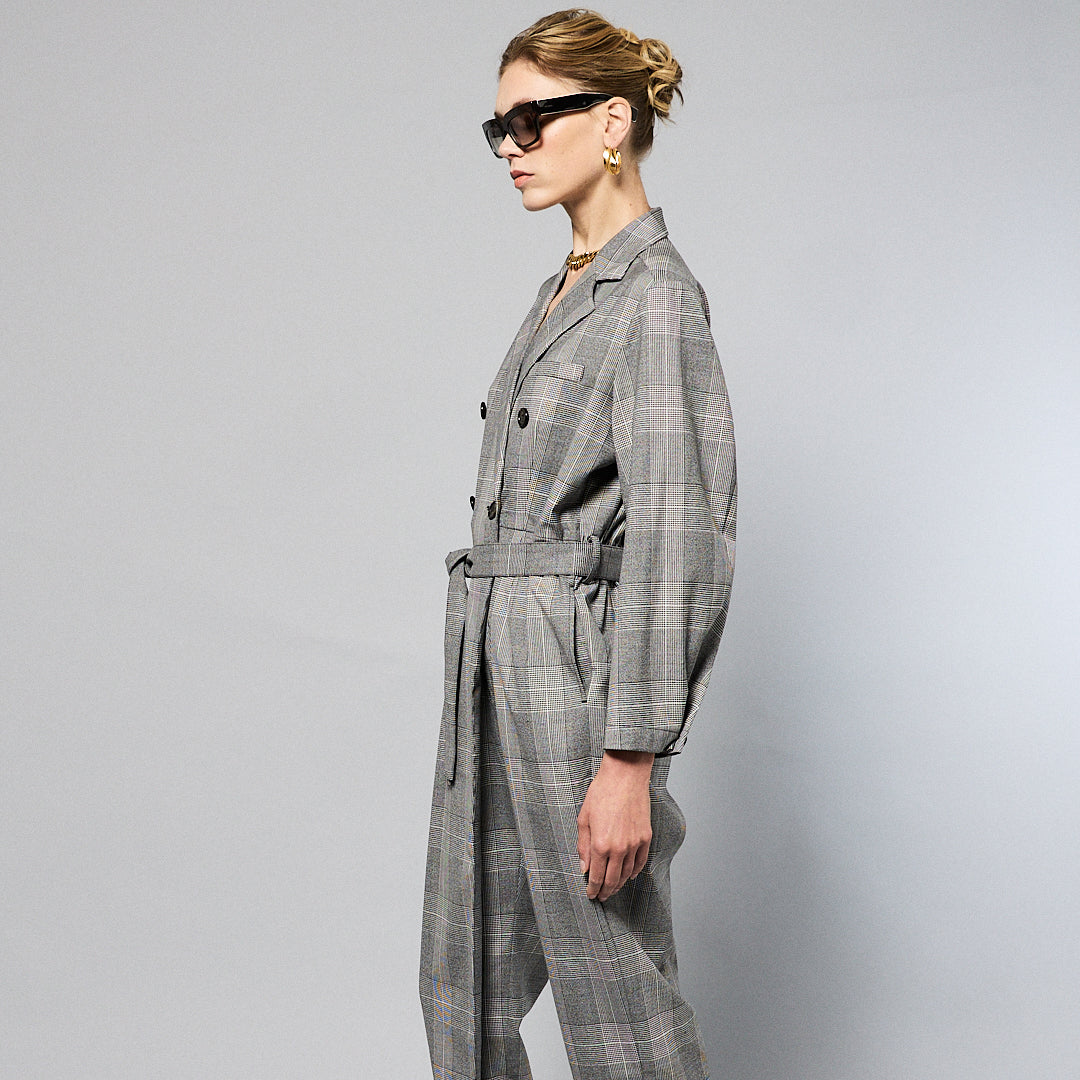 A woman stands against a plain background wearing a Chloe Glen Plaid Tailored jumpsuit by Maison Lilli. She accessorizes with large black sunglasses, gold hoop earrings, and a gold necklace. Her hair is styled in an updo.