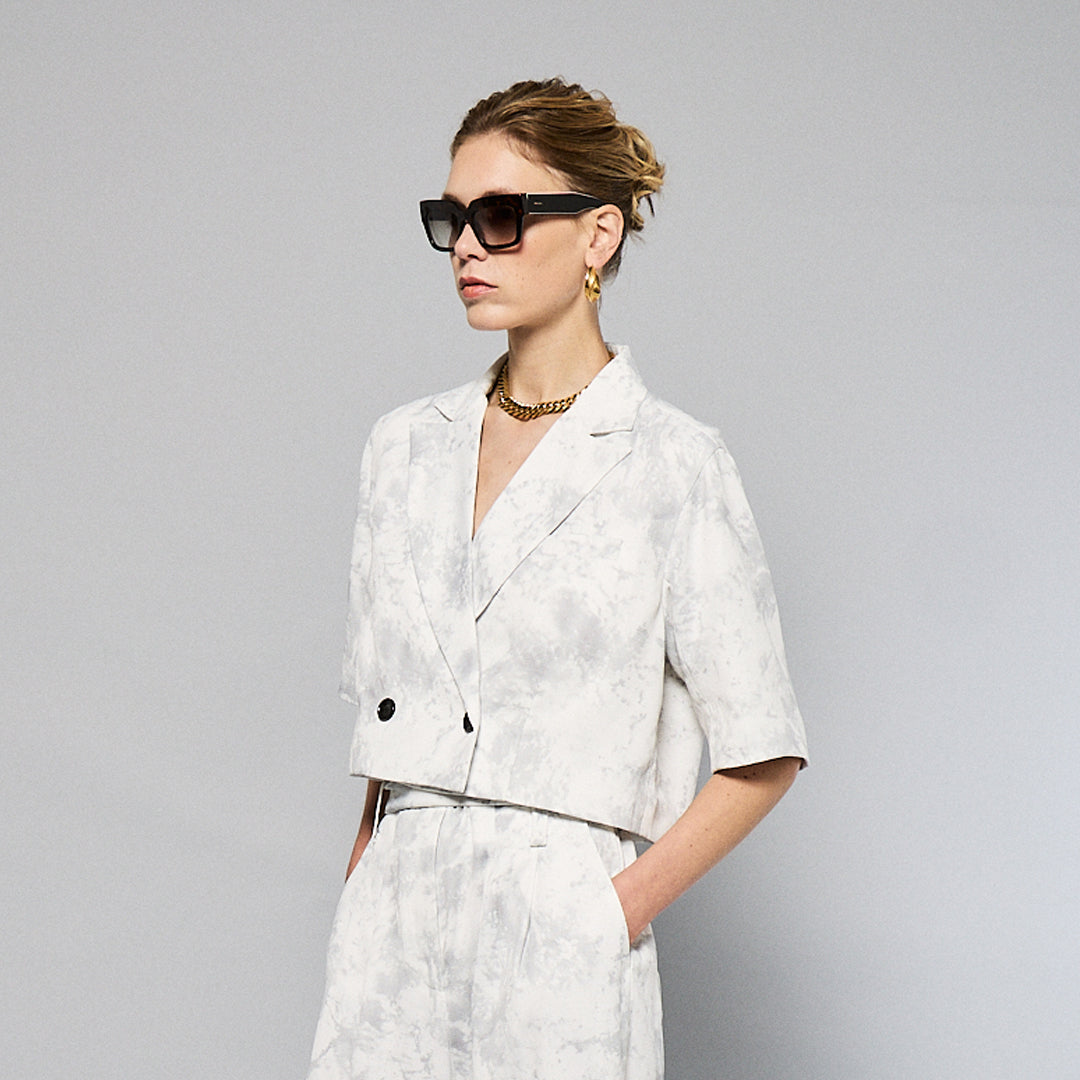 A person stands against a plain background, wearing a Marble Print Short Sleeve Cropped Jacket by Maison Lilli. She also wear a gold hoop earrings, and a thick gold chain necklace.