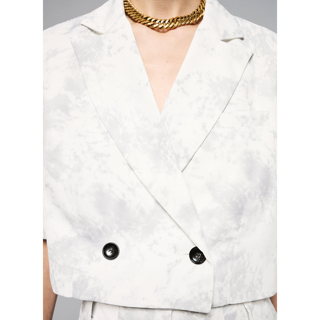 A person stands against a plain background, wearing a Marble Print Short Sleeve Cropped Jacket by Maison Lilli. She also wear a gold hoop earrings, and a thick gold chain necklace.