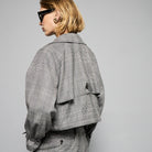A person with short blond hair wearing oversized black sunglasses, a Marie Cropped Glen Plaid Jacket by Maison Lilli in a soft wool blend with an adjustable back waist, a beige shirt, and a gold chain necklace stands against a neutral gray background. The individual exudes a confident and stylish demeanor.