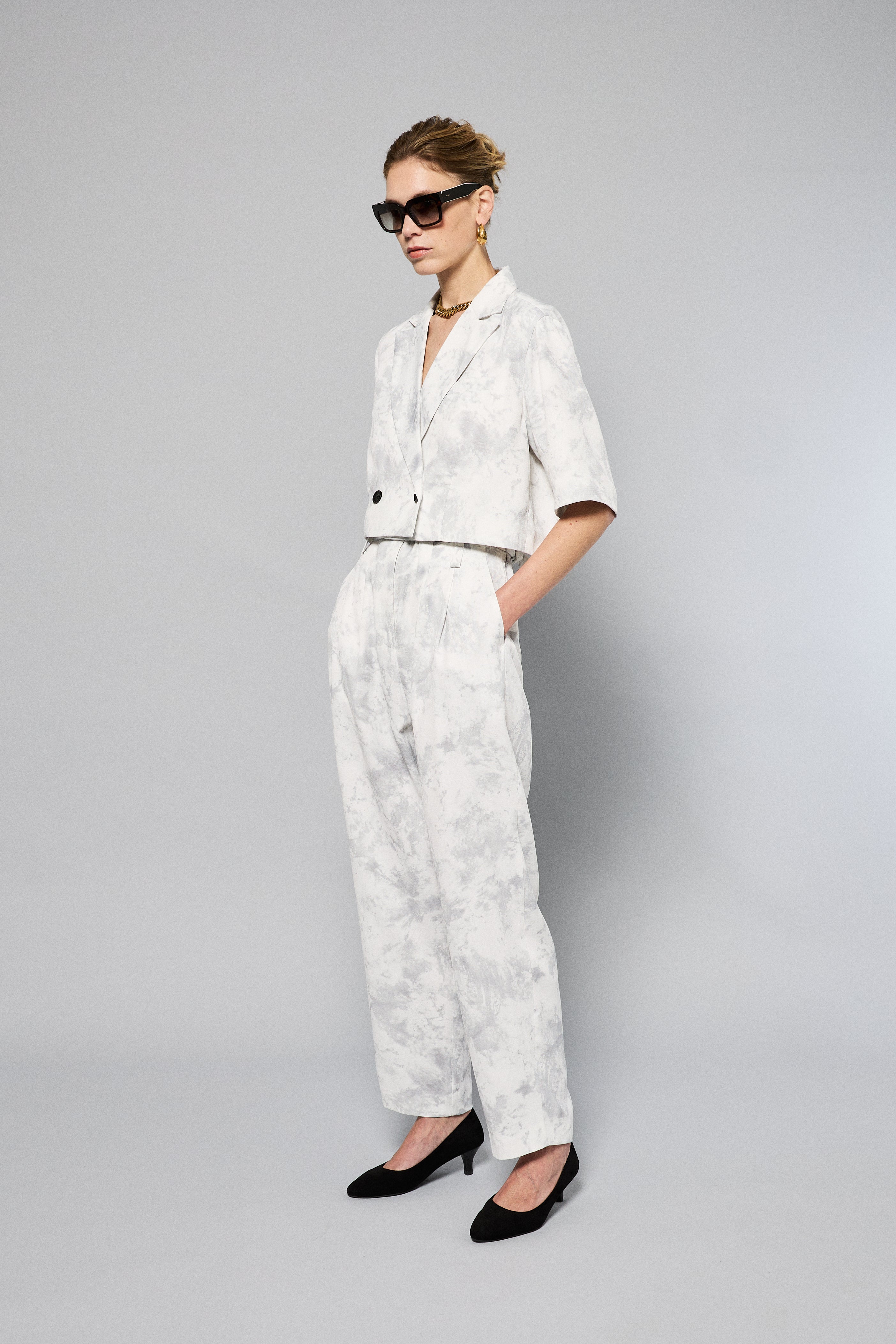 A person is wearing Maison Lilli Paperbag Pleated Pants - Marble Print. The pants have pockets and a removable belt. The person also sports a matching top and black high-heeled shoes against a plain light gray background.