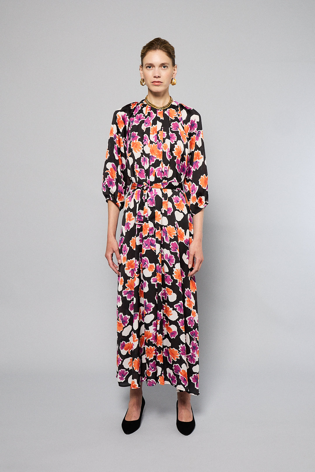 A woman stands against a plain background wearing the Maison Lilli Nathalie Printed Maxi Dress featuring a vibrant floral pattern with shades of pink, orange, and white on a black base. She has her hair pulled back and is accessorizing with gold earrings.