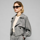 A person with short blond hair wearing oversized black sunglasses, a Marie Cropped Glen Plaid Jacket by Maison Lilli in a soft wool blend with an adjustable back waist, a beige shirt, and a gold chain necklace stands against a neutral gray background. The individual exudes a confident and stylish demeanor.
