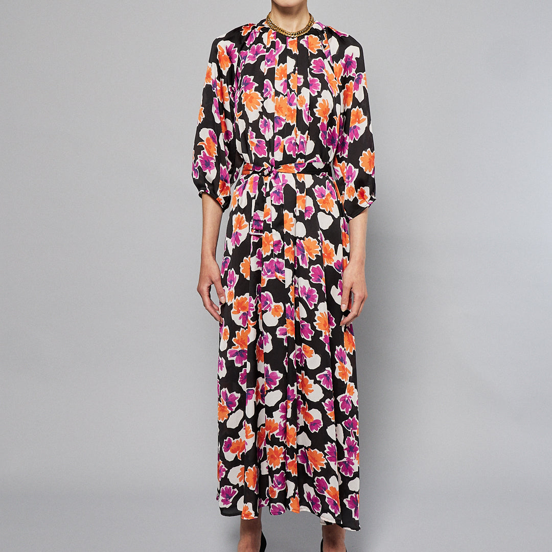 A woman stands against a plain background wearing the Maison Lilli Nathalie Printed Maxi Dress featuring a vibrant floral pattern with shades of pink, orange, and white on a black base. She has her hair pulled back and is accessorizing with gold earrings.