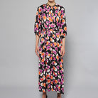 A woman stands against a plain background wearing the Maison Lilli Nathalie Printed Maxi Dress featuring a vibrant floral pattern with shades of pink, orange, and white on a black base. She has her hair pulled back and is accessorizing with gold earrings.