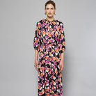 A woman stands against a plain background wearing the Maison Lilli Nathalie Printed Maxi Dress featuring a vibrant floral pattern with shades of pink, orange, and white on a black base. She has her hair pulled back and is accessorizing with gold earrings.