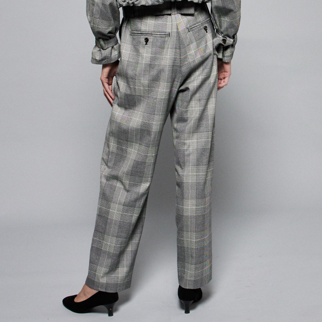 A person is wearing high-waisted, gray wool-blend Paperbag Pleated Pants - Glen Plaid by Maison Lilli with a large belt buckle. She also wearing a loose-fitting short-sleeved off-white top and black heels. The background is plain light gray.
