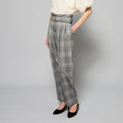 A person is wearing high-waisted, gray wool-blend Paperbag Pleated Pants - Glen Plaid by Maison Lilli with a large belt buckle. They have one hand in their pocket and are also wearing a loose-fitting short-sleeved off-white top and black heels. The background is plain light gray.