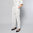 A person is wearing Maison Lilli Paperbag Pleated Pants - Marble Print. The pants have pockets and a removable belt. The person also sports a matching top and black high-heeled shoes against a plain light gray background.