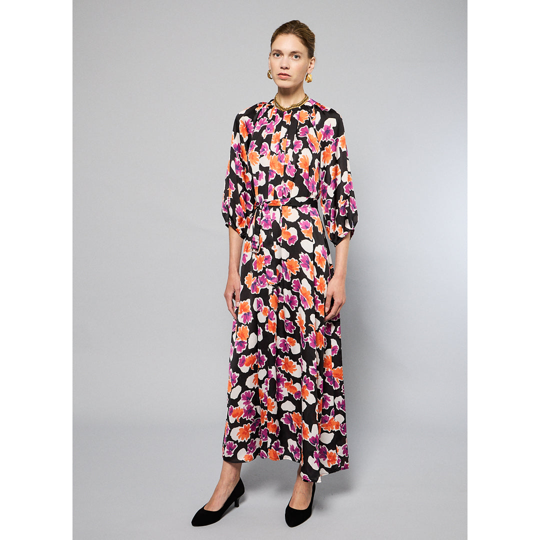 A woman stands against a plain background wearing the Maison Lilli Nathalie Printed Maxi Dress featuring a vibrant floral pattern with shades of pink, orange, and white on a black base. She has her hair pulled back and is accessorizing with gold earrings.