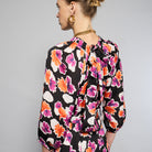 A woman stands against a plain background wearing the Maison Lilli Nathalie Printed Maxi Dress featuring a vibrant floral pattern with shades of pink, orange, and white on a black base. She has her hair pulled back and is accessorizing with gold earrings.