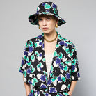 A person stands against a plain background, wearing a bucket hat and matching Yelena Short Sleeve Cropped Jacket - Floral Print by Maison Lilli in blue, green, and white. They also wear a white top underneath, gold hoop earrings, and a thick gold chain necklace.