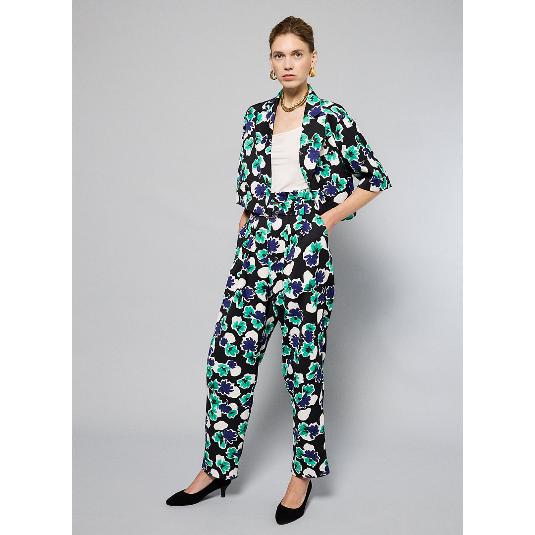 A person is wearing Maison Lilli Paperbag Pleated Pants - Floral Print with large blue, green, and white flowers. The pants have pockets and a removable belt. The person also sports a matching top and black high-heeled shoes against a plain light gray background.