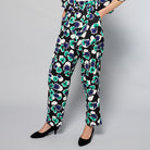 A person is wearing Maison Lilli Paperbag Pleated Pants - Floral Print with large blue, green, and white flowers. The pants have pockets and a removable belt. The person also sports a matching top and black high-heeled shoes against a plain light gray background.