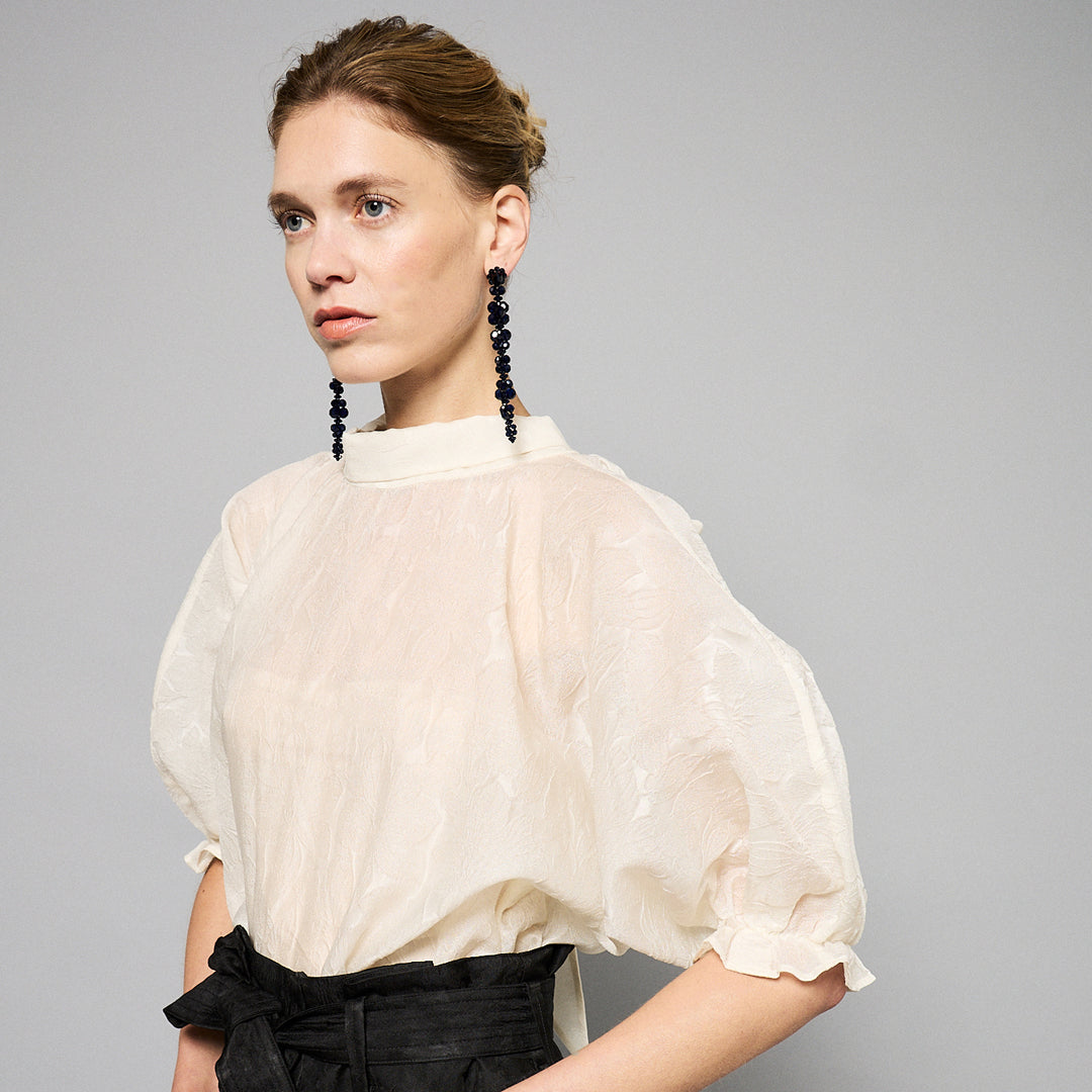 A woman with light brown hair wearing a delicate, translucent Maison Lilli Amelie Sheer Jacquard Top and black high-waisted pants, paired with long, black beaded earrings, posing against a soft gray background.