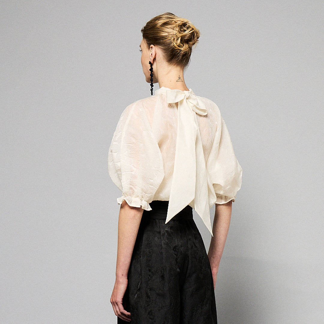 A woman with light brown hair wearing a delicate, translucent Maison Lilli Amelie Sheer Jacquard Top and black high-waisted pants, paired with long, black beaded earrings, posing against a soft gray background.