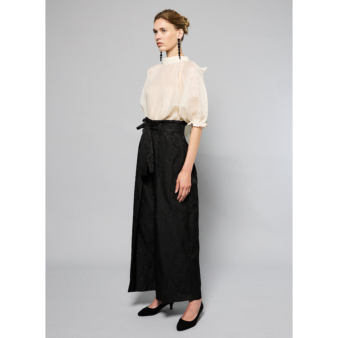 A woman with light brown hair wearing a delicate, translucent Maison Lilli Amelie Sheer Jacquard Top and black high-waisted pants, paired with long, black beaded earrings, posing against a soft gray background.