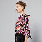 A woman wearing a Maison Lilli Amelie Floral Printed Top with vibrant pink and orange flowers and black pants stands against a grey background. Her hair is styled back, and she wears large, round earrings.