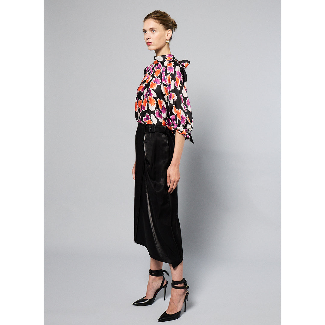 A woman wearing a Maison Lilli Amelie Floral Printed Top with vibrant pink and orange flowers and black pants stands against a grey background. Her hair is styled back, and she wears large, round earrings.