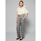 A person is wearing high-waisted, gray wool-blend Paperbag Pleated Pants - Glen Plaid by Maison Lilli with a large belt buckle. They have one hand in their pocket and are also wearing a loose-fitting short-sleeved off-white top and black heels. The background is plain light gray.