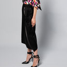 A person standing in front of a plain gray background, wearing a colorful floral blouse, a Maison Lilli Jodie Sparkling Satin Asymmetrical Draped Skirt, and black strappy high heels. The shot captures their lower torso and legs.