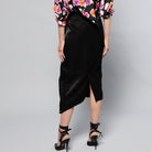A person standing in front of a plain gray background, wearing a colorful floral blouse, a Maison Lilli Jodie Sparkling Satin Asymmetrical Draped Skirt, and black strappy high heels. The shot captures their lower torso and legs.