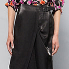 A person standing in front of a plain gray background, wearing a colorful floral blouse, a Maison Lilli Jodie Sparkling Satin Asymmetrical Draped Skirt, and black strappy high heels. The shot captures their lower torso.